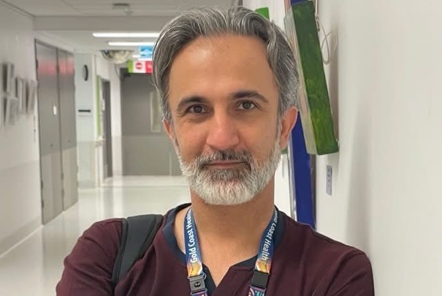 A man with a greying beard and a maroon shirt leaning against a white wall.