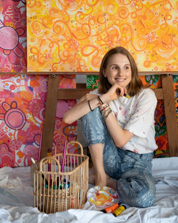 Artist smiling with paint supplies sitting in front of canvas