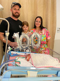 Tillie sleeping in a cot with dad Anthony, sister Kobie and mum Ashleigh standing behind holding an inflated balloon shaped as the number 100.